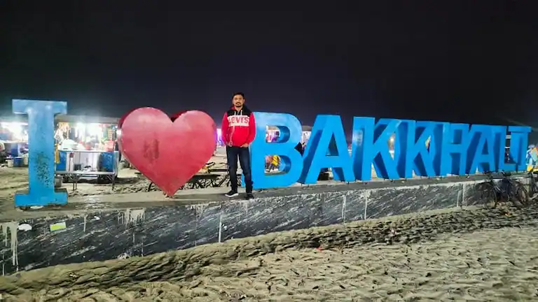 Tourist standing at the illuminated I Love Bakkhali signboard at night.