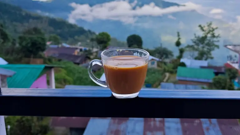 Glass cup of hot tea on a homestay balcony railing overlooking Icche Gaon village and misty green mountains.