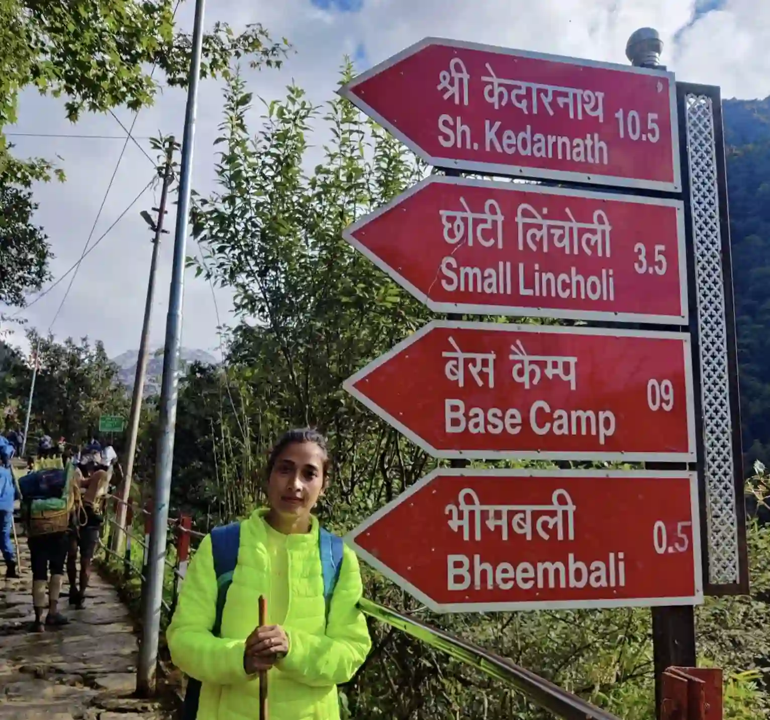 A female trekker standing next to a red government signboard showing remaining walking distances to Bheembali, Lincholi, and Base Camp.