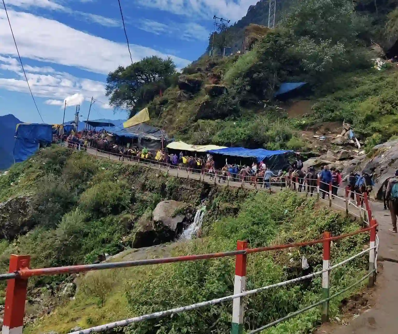 Pilgrims and mules climbing a steep, winding mountain path lined with safety railings and small stalls.
